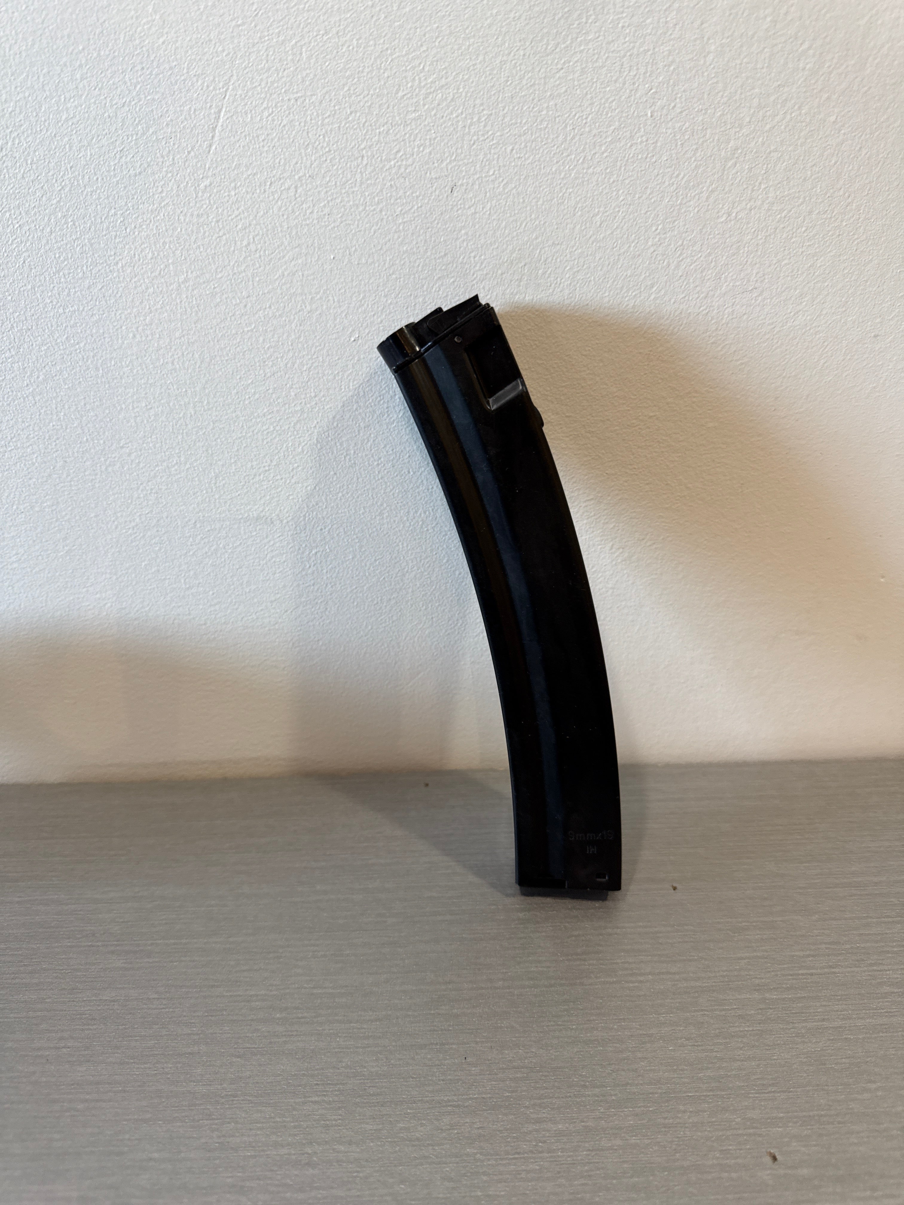 Black curved used MP5 airsoft magazine standing upright on tabletop against white wall