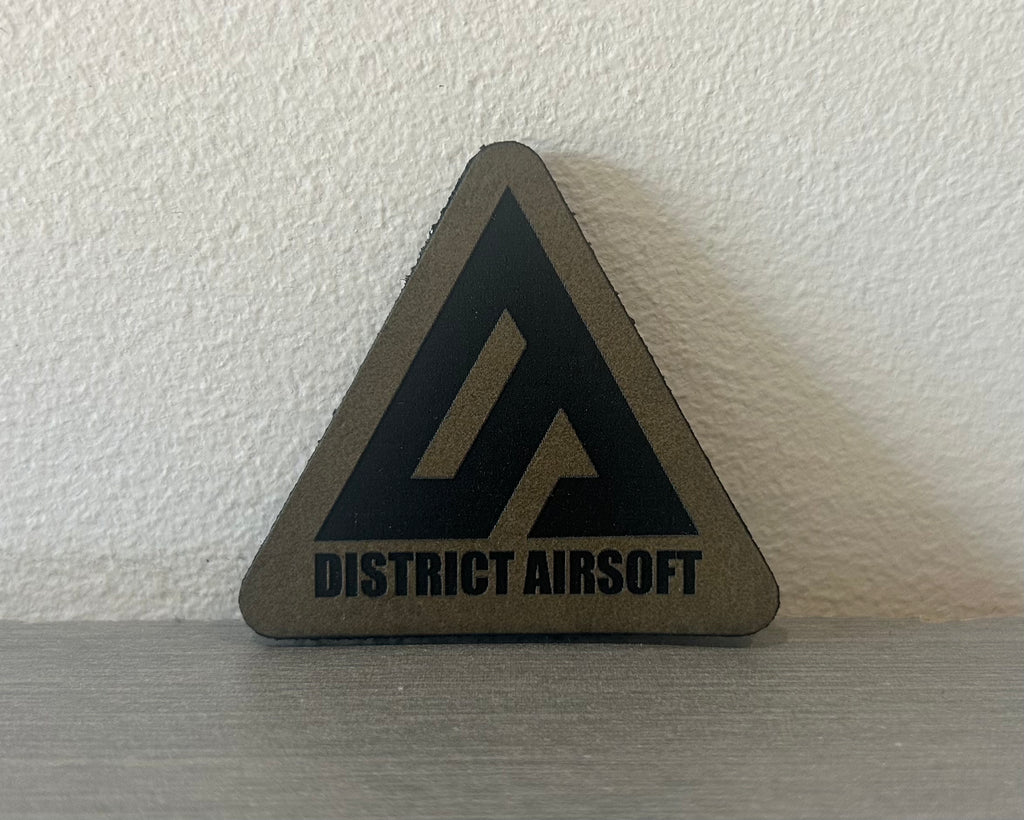 District Airsoft Leather Patch in a triangular shape with a black logo on a gray background