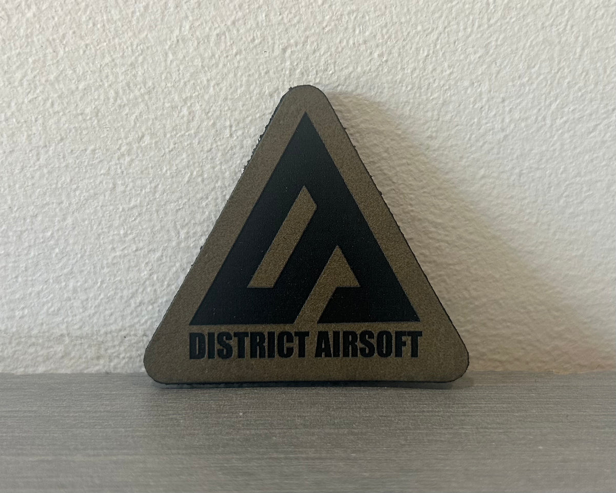 District Airsoft Leather Patch in a triangular shape with a black logo on a gray background