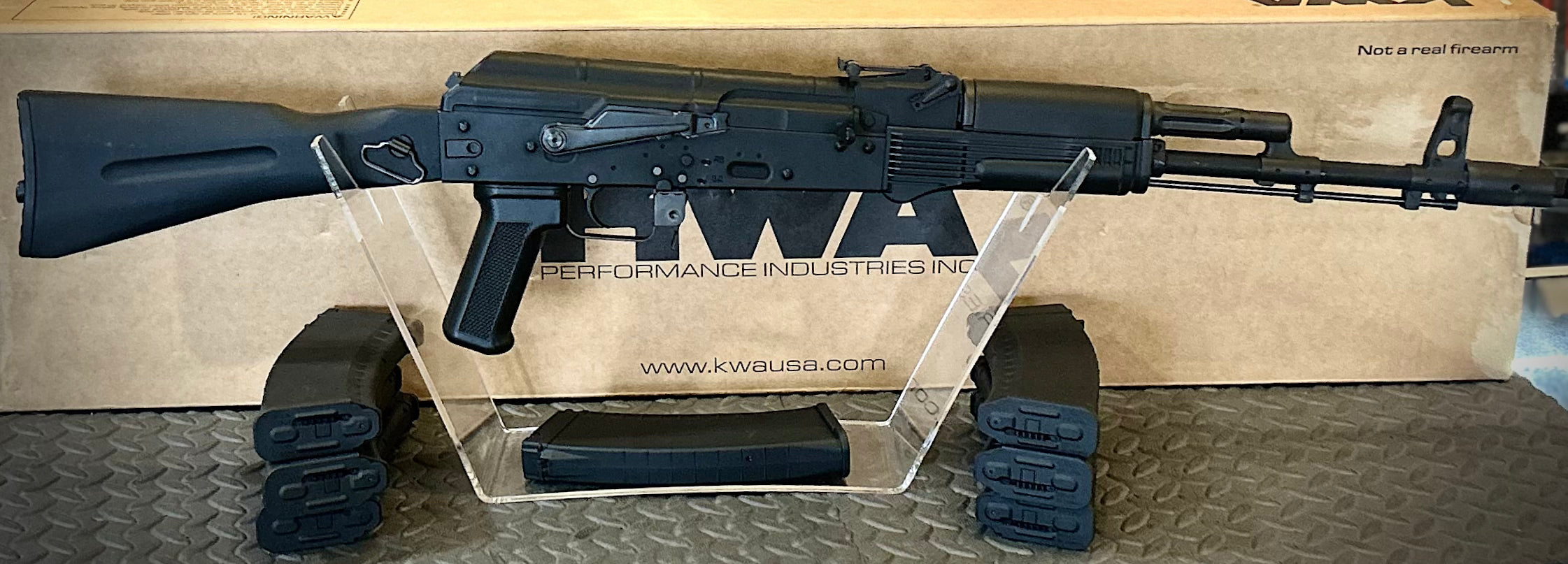 KWA AK 74M airsoft rifle displayed with magazines on a stand and branded packaging in the background